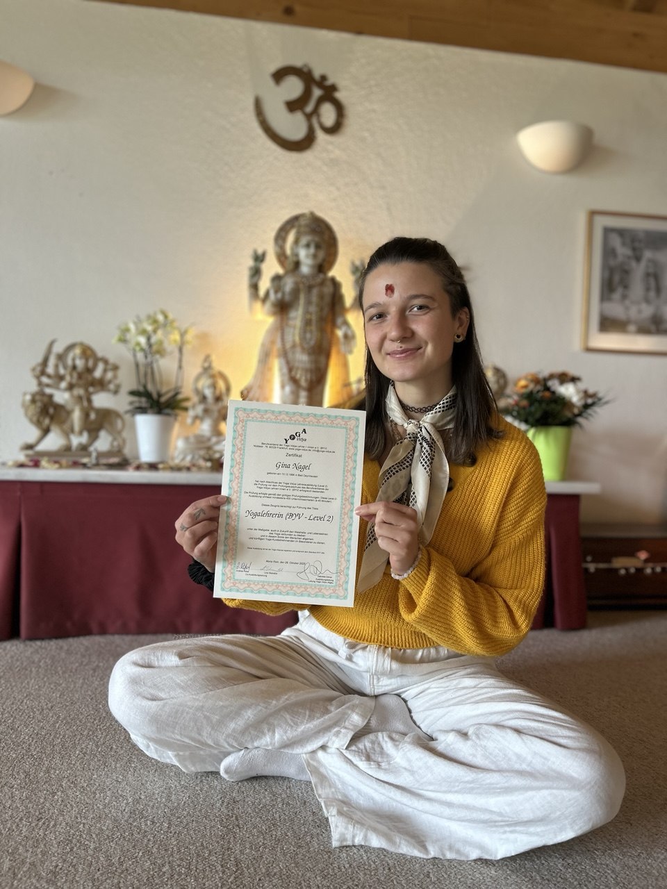Gina Nagel – zertifizierte Yogalehrerin (BYV Level 2), sitzend im Schneidersitz mit ihrem Zertifikat in einem schönen Yogaraum